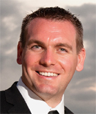 A smiling man with short brown hair wearing a black suit and white shirt against a cloudy sky.