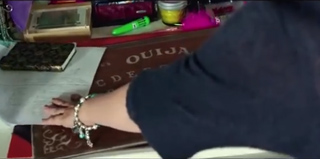 A person's hand with bracelets rests on a brown Ouija board on a table.