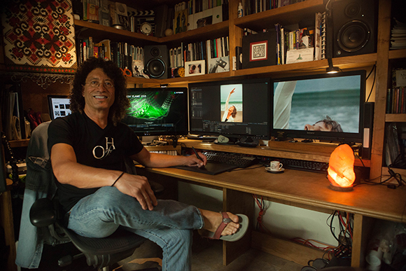 A man sits in a chair at a wooden desk with three computer monitors and various studio equipment.