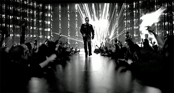 A man in dark clothing walks down a glowing runway toward a crowd of people with raised hands.