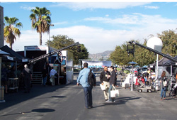 Outdoor street scene with palm trees, tents, and film cranes set up on both sides of the road.