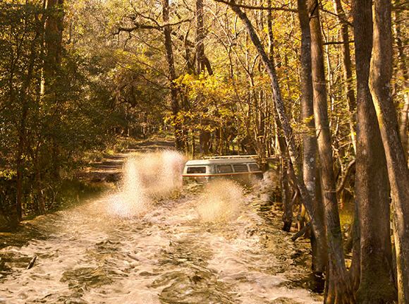 A white van splashes through a shallow, flowing stream in a sunlit forest.