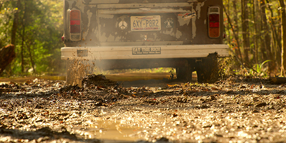 Rear view of a weathered truck driving through deep mud on a wooded trail.