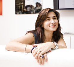 A smiling woman with long brown hair leans her arms on a white surface, wearing several bracelets and rings.