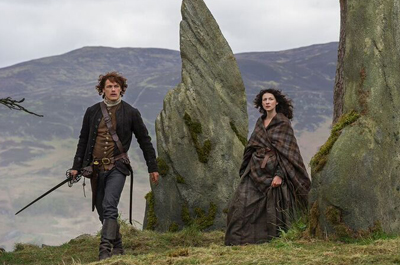 A man in a dark jacket and sword walks near a woman wrapped in a plaid shawl by large mossy stones.