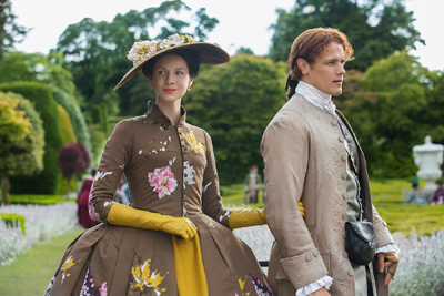 A woman in a floral brown gown and a man in a tan frock coat walk through a lush green garden.