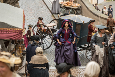 A woman in a purple period dress walks through a busy cobblestone street with horse-drawn carriages.