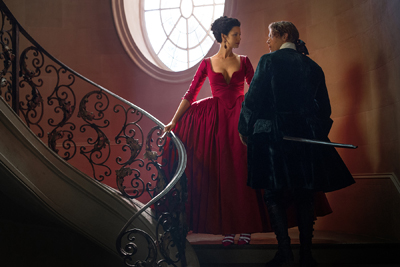 A woman in a long red gown stands on a curved staircase facing a person in dark attire.