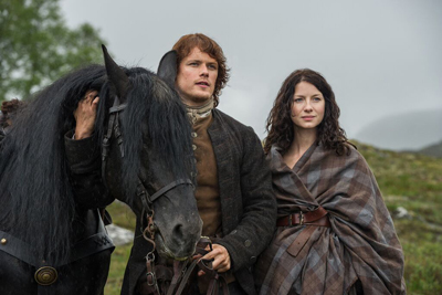 A man in period clothing stands by a black horse next to a woman wrapped in a plaid shawl in a misty field.