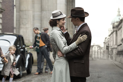 A man and woman in 1940s clothing embrace on a cobblestone street near a large building.