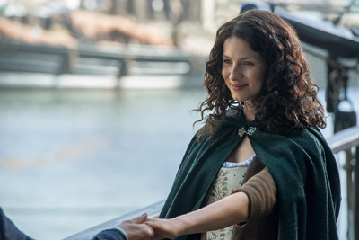 A woman with long curly hair and a dark green cloak smiles while holding someone's hand.