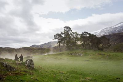 Two people sit on a grassy hill amidst low-lying mist with trees and snow-capped mountains behind them.