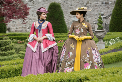 Two women in elaborate 18th-century gowns and hats standing in a manicured garden.