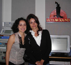 Two women smiling and posing together in a music studio with recording equipment and a logo on the wall.