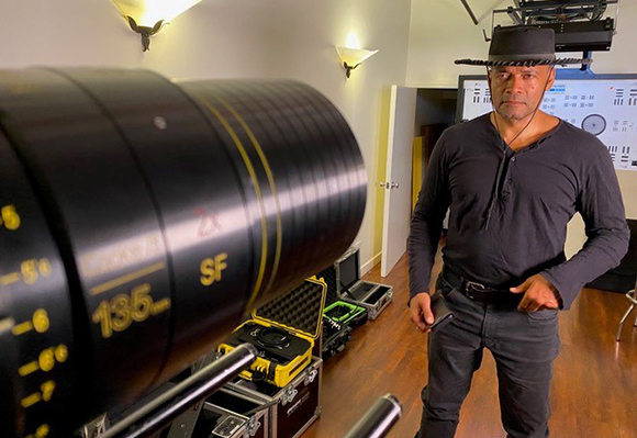 A man in a black shirt and hat stands behind a large, close-up cinema lens in a studio.