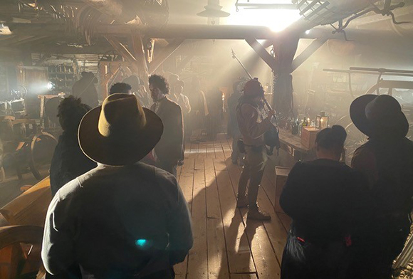People in period clothing stand in a dusty, sunlit wooden structure with beams and planks.