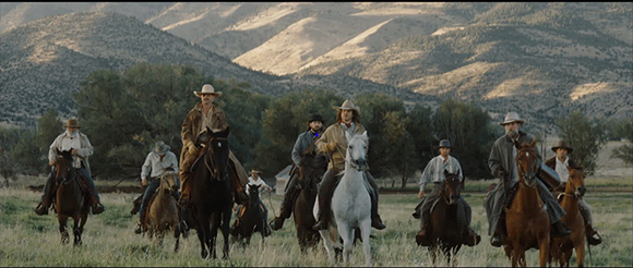 A group of men on horseback riding through a field toward the viewer with hills in the background.