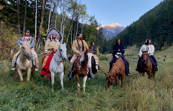 People in traditional Indigenous clothing riding horses through a grassy mountain valley.