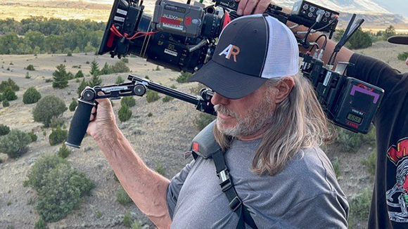 A man with long hair and a grey cap holds a large professional cinema camera outdoors.