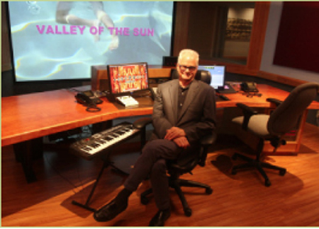 A man in a suit sits at a large wooden desk in a studio with a screen displaying 'VALLEY OF THE SUN'.