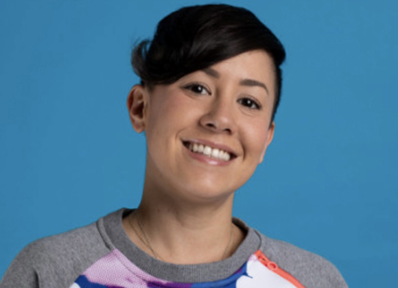 A person with short dark hair smiling against a solid blue background.