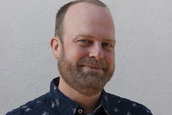 A man with short light hair and a brown beard smiling against a plain light wall.