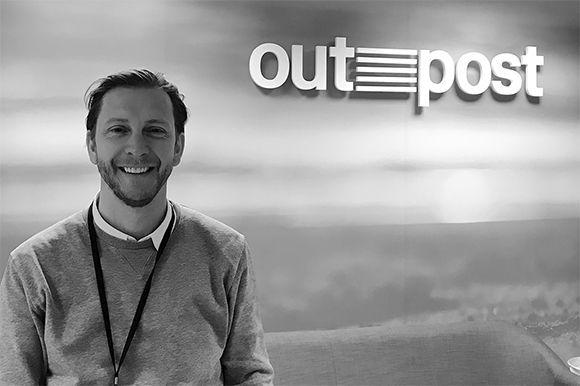 A smiling man in a sweater stands in front of a wall with the word 'outpost' in large white letters.