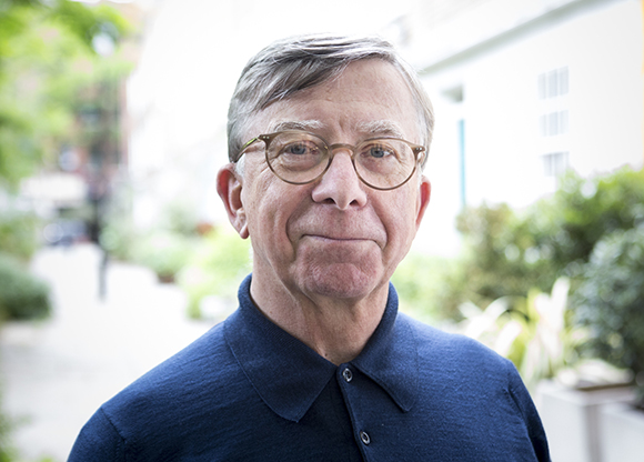 A man with short brown hair and round glasses wearing a dark blue polo shirt outdoors.