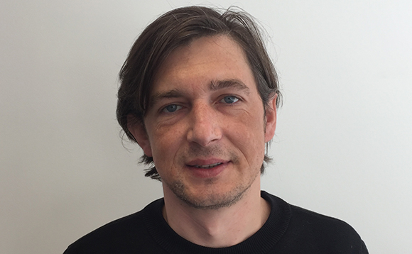 A man with short brown hair and blue eyes wearing a black sweater against a white background.