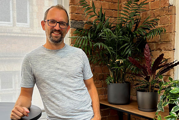 A man with glasses and a beard wearing a grey t-shirt stands near potted plants against a brick wall.
