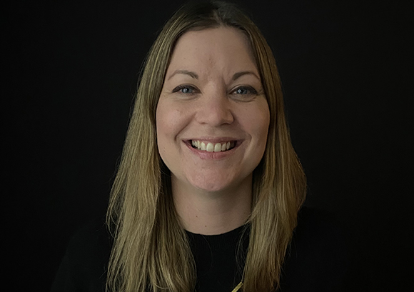 A smiling woman with long blonde hair looking directly at the camera against a solid black background.