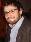A man with dark hair, glasses, and a beard wearing a light blue shirt and a dark jacket.