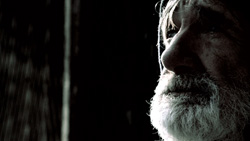 Close-up profile of an elderly man with white hair and a thick white beard against a dark background.