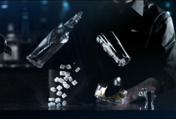 A bartender pours liquid from a bottle and shaker over falling sugar cubes in a dark setting.