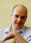 A smiling man with a receding hairline wearing a light blue striped collared shirt.