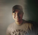 A smiling man wearing a dark baseball cap and a grey t-shirt with text on it.