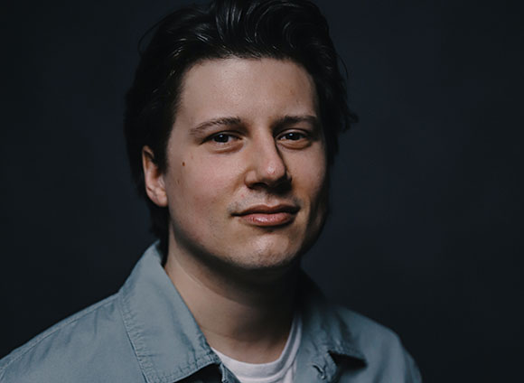 A man with dark hair and a slight smile looks toward the camera against a dark background.