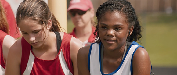 Two young female athletes in red and white athletic wear sit outdoors near a person in a red cap.