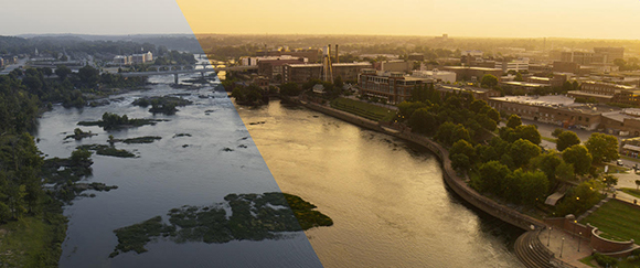 Aerial view of a wide river winding through a city with buildings and trees under a golden sunset sky.