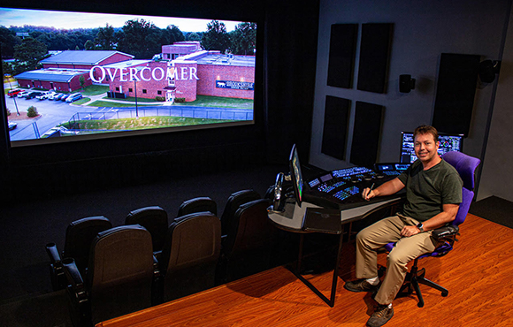 A man sits at a technical control desk in a dark theater facing a large screen displaying a movie title.
