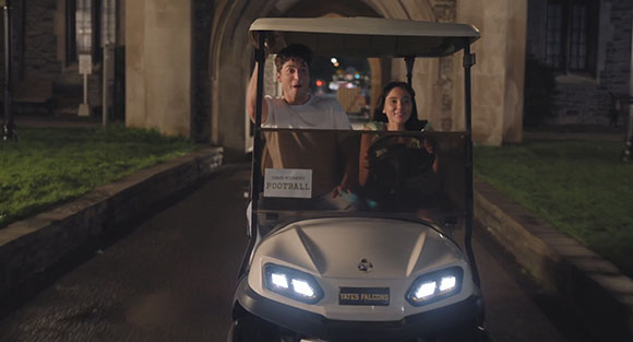 A man and a woman smiling while riding in a white golf cart down a paved path at night.