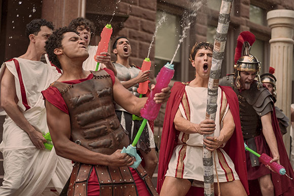 People dressed in Roman attire spraying water from colorful water guns outdoors.