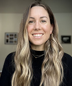 A smiling woman with long, wavy blonde hair wearing a black turtleneck.
