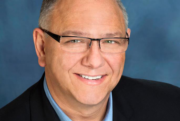 A smiling man with short graying hair and rectangular glasses wearing a dark blazer and light blue shirt.