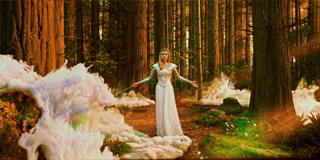 A woman in a long white dress stands with arms outstretched in a dense redwood forest with swirling white mist.