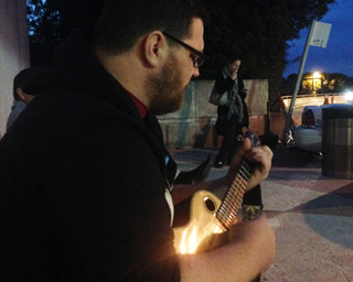 Man with beard and glasses plays a hollow-body electric guitar outdoors at dusk. Another person stands in the background.