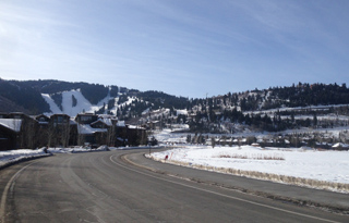 A paved road curves through a snowy landscape towards a ski resort with slopes visible on the mountainside.