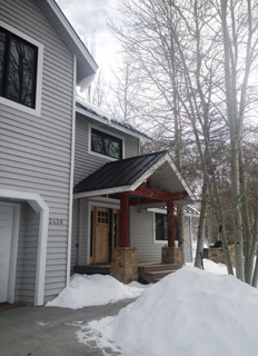 Exterior of a light gray house with a dark roof, wooden front door, and stone pillars on the porch, surrounded by snow and ba