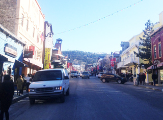 A street lined with shops and buildings in a mountain town, with cars driving and people walking on a sunny winter day.