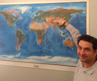 A man in a collared shirt points to a large, detailed world map displayed on a wall.